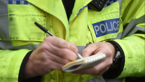 A close-up image of a police officer's chest. He is wearing a high-visibility police jacket and writing in a notebook