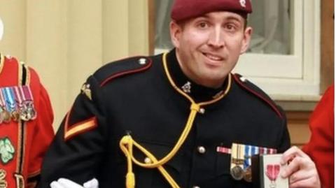 An image of paratrooper Ben Parkinson from the chest up. He is in uniform, a black jacket with red trim on the epaulettes on each shoulder and with a gold rope trailed across his chest, and a burgundy beret on his head. He has medals pinned on his uniform and is holding an MBE. 