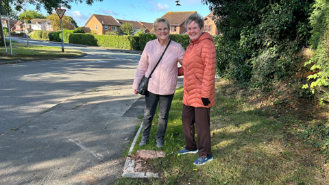 Two women standing next to a concrete plinth where a post box was sited before it was stolen, with a road to their right and trees to their left