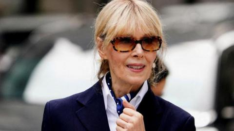 E Jean Carroll with a navy blazer, white shirt and blue patterned scarf. She is wearing large sunglasses and looking straight ahead. 