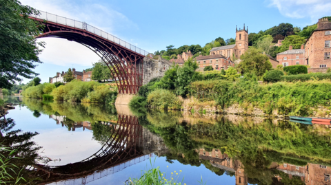A small town with red brick buildings, a church and a metal framed bridge reflected in a still river
