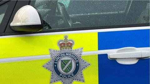 The side of a Lincolnshire Police patrol car. It has blue and yellow fluorescent markings and the force's badge on the door