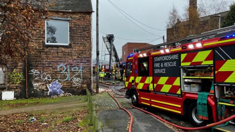 A fire engine in the foreground can be seen with hoses trailing alongside it, with further fire engines and firefighters in the background and a ladder is being raised.