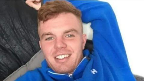 Jack O'Brien, wearing a royal blue fleece long-sleeved top, smiles at the camera as he relaxes on a sofa.