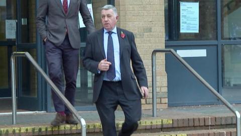 A man, with short grey hair, is leaving the magistrates' court. He is wearing a dark blue suit, with light blue shirt and dark blue tie. He has a poppy on his left lapel