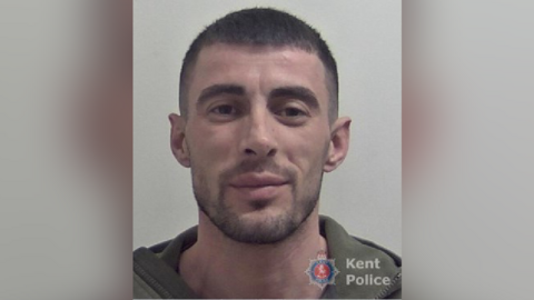 A police mugshot of a smirking man with short brown hair and stubble. 
