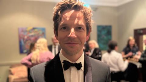 Damien Molony dressed in a formal black tuxedo with a white dress shirt and a black bow tie stands indoors at what appears to be a social or formal event. The background is softly lit and out of focus, showing several people mingling, artwork on the walls, and glasses set out on a table. 