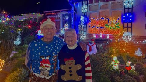 Bill and Barbara wearing Christmas jumpers, smiling outside their house which is covered in Christmas lights and illuminated objects including snowmen and Santa dogs.