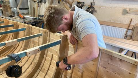 A bearded man with ginger hair in a grey polo shirt is leaning over the side of a wooden boat he is building and drilling a nail into one of its sides in his workshop.