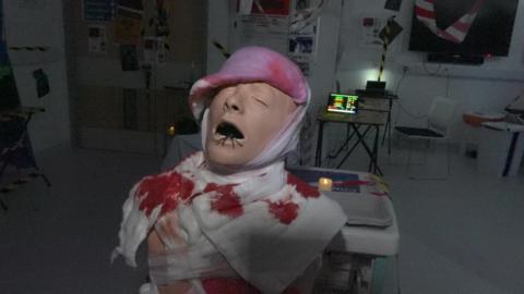 A mannequin covered in bloodstained bandages. It is Caucasian and has its mouth open. A spider is crawling out. It is in a darkened room with artificial candles lighting up the room. Behind is a monitor screen on a desk with an empty chair nearby.