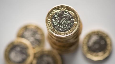 Close up image of a selection of British pound coins. The coins are stacked on top of one another. A couple of £1 coins are scattered around the stack. 