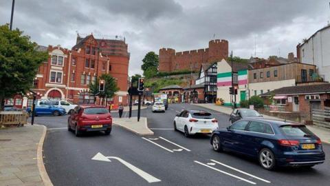 General view of Shrewsbury station gyratory - a number of cars on the road with a large historic red brick building and castle in the background of the photo