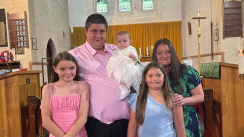 Dad Shaine Kirby holding baby Paighton in the church with mum Elizabeth Lambert and siblings Lexi and Erin by his side