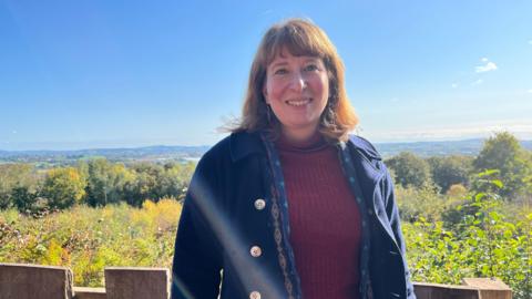 A woman with shoulder length brown hair is smiling. She is wearing a red turtle neck top underneath a blue  jacket with gold buttons. There are trees and fields in the background