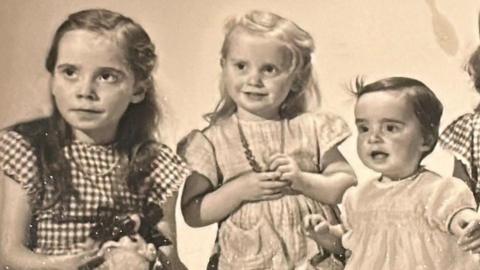 Old yellowing black and white photo of the three sisters as young children. The eldest on the left is wearing a checked dress and has long brown hair. The middle girl is younger and has blond hair and is wearing a plain dress while holding a necklace around her neck. The youngest on the right is a toddler with short dark hair.
