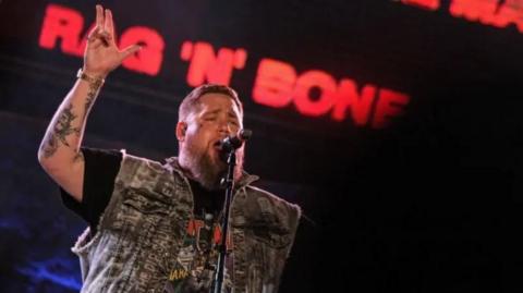 Rag'n'Bone Man performing on stage with a microphone on a stand in front of him and the words 'Rag'n'Bone Man' in red writing on a black stage background behind him.