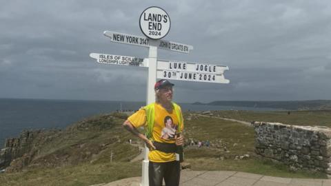 A young man in running clothes stands at a large white sign holds a bottle of wine. He is near the edge of cliffs over the sea,