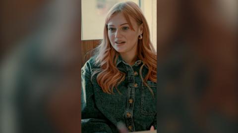 A woman with long wavy pink hair is talking to someone off camera. She is wearing a denim jacket with buttons.