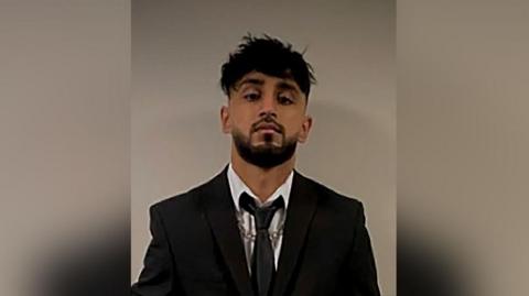 Uzair Khan with black hair and beard wearing a black suit and tie with a silver chain necklace.