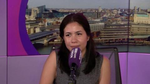 A woman speaks into a microphone against a backdrop of a city skyline and purple.
