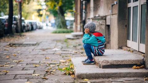 A young child wearing a striped blue raincoat and grey hat sitting on a concrete step outside a building on a deserted street, holding their head in their hands