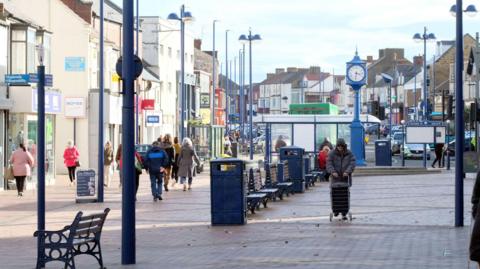 A high street with shoppers walking up and down a pedestrianised road. Benches and bins are lining the centre of the street. A blue town clock is next to a bus shelter in the distance.