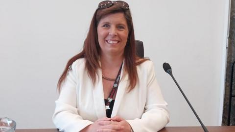 Ceri Cawley is smiling at the camera with a white suit jacket on and sunglasses on top of her head. A microphone is positioned in front of her and a water jug is in the bottom left of the picture.