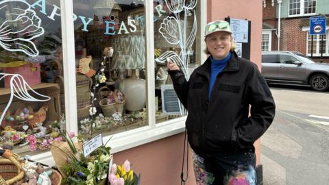 Juliet is wearing a cap and a dark coat.
She is standing next to a shop with a white, floral window mural that she painted on it.
The shop is on the High Street and there are flowers next to it.