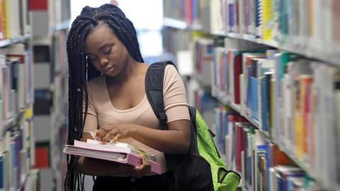 A young university sudent looks through books as she stands between bookshelves in a university library