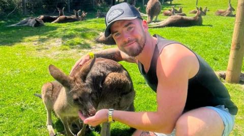 Joe O'Sullivan is on the left of picture, wearing a baseball cap and a black singlet over light blue shorts. He is hand-feeding a kangaroo.