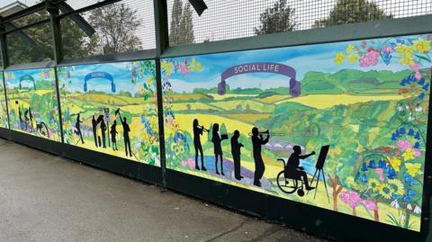 A train bridge with a mural displaying rural scenes, flowers and silhouette figures of children