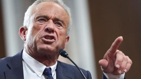 RFK Jr speaks into a microphone while raising his left hand and pointing his index finger toward the senate committee