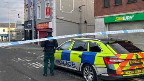 A police officer stands with his back to the camera and is stood beside a police car. There is a police cordon closing off the street. In the distance there is the Lurgan police station.