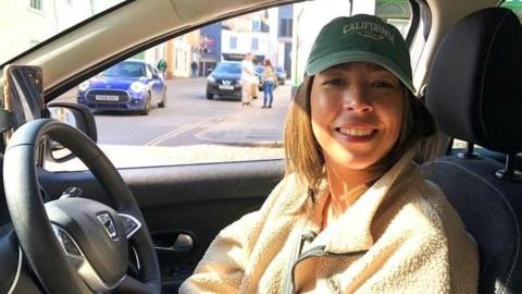 A woman Sabrina Pettit is sitting in her taxi in the drivers seat. She is wearing a beige fleece and brown baseball cap. Sabrina is smiling at the camera. It's a sunny day outside and you can see a street view out the window behind her. 