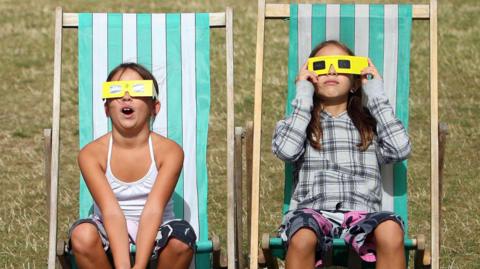 Two girls are sitting on green and white stripy deck chairs on grass wearing yellow eclipse glasses. They're looking up and it is a bright day.