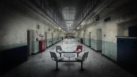 Four chairs and a small table are placed in the long hallway of Shepton Mallet prison. There is a net which separates the ground floor with the other floors.
