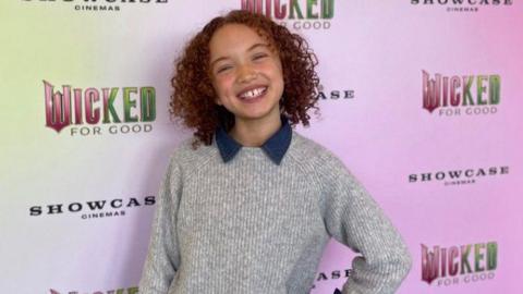 Esme smiling at the camera as she attends the screening, wearing a grey jumper with a blue collar. She has brown curly hair.