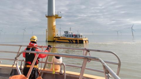 An engineer standing on the deck of a boat wearing orange and black clothing as another boat approaches one of a number of offshore wind turbines.