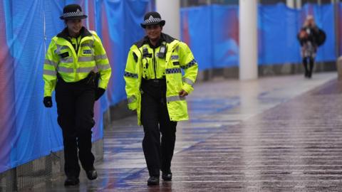 Two female police officers patrolling a wet street in the UK. The officers are both wearing high viz police coats with black trousers, black boots and a black police hat.