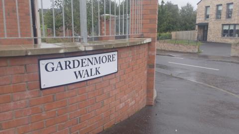 A red brick wall with a silver railing on top of it with a white and black road sign with 'Gardenmore Walk' written on it.