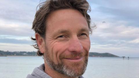 A picture of Greg smiling at the camera. He has brown hair and a short grey and brown beard.