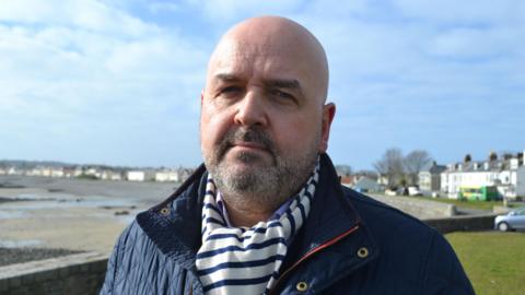 Jonathan Le Tocq is dressed in a blue jacket with a blue and white striped scarf. There are houses in the background around a bay, with the beach on the left of the picture. The tide is out.