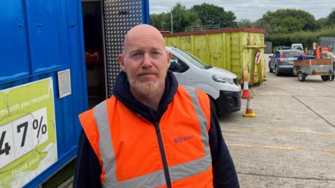 Simon Collett is pictured in a navy hoodie with a high-vis vest over it. He has a beard and a bald head and his expression is serious. He is standing in front of metal containers for various types of waste at a waste and recycling plant.