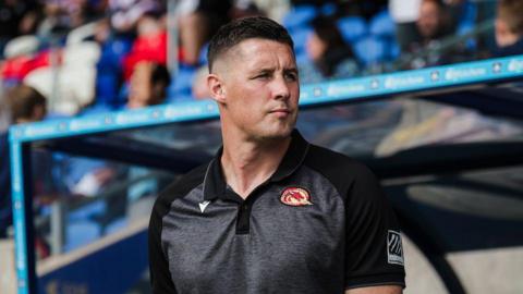 Sam Tomkins standing in the dugout looking over his left shoulder during a game for Catalans Dragons in 2025