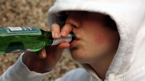 A boy wearing a white hoodie which covers half of his face. He is drinking from a green bottle of lager.