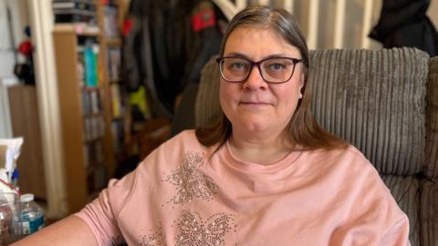 A woman with long brown hair is sitting in a chair wearing glasses and a pink top with butterflies made from gems on it. The chair is grey corduroy and on a table next to her is a plastic bottle of water and a glass