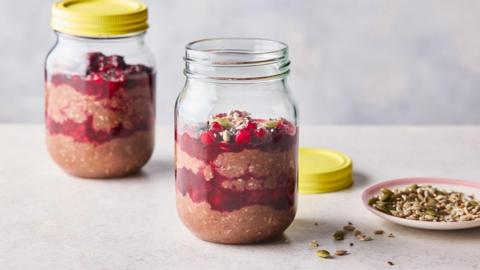Two jars filled with chocolate oats and berry compote, topped with a handful of berries. One of the jars is closed with a yellow lid while the other is open, with the lid on the countertop