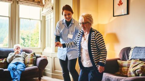 Care worker in a pale blue uniform helps an elderly woman in a navy and white striped cardigan up from a chair in a care home. A man sits on a sofa under a window in the background. 