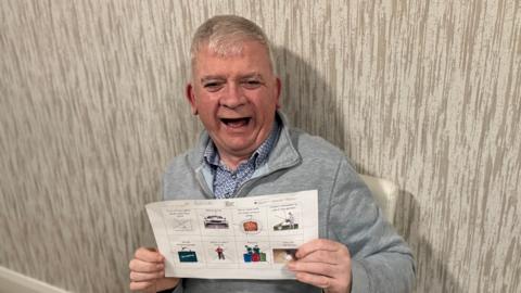 A grey haired man with a grey half zip holds up a sheet with words and pictures on it. He is smiling and happy. 