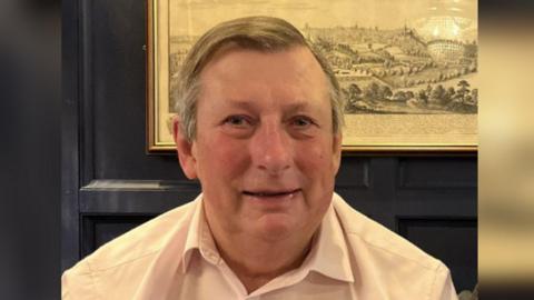 A man with grey hair wearing a white shirt smiles at the camera. He is sitting in a room with black panelling and an old-fashioned drawing on the wall.